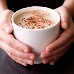 Hands hold a white mug filled with frothy coffee, sprinkled with cocoa, against a dark surface.
