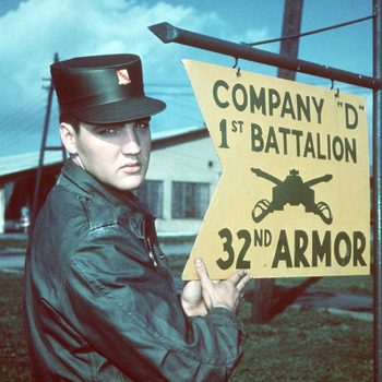 Man holding a sign for Company "D" 1st Battalion 32nd Armor, outdoors near military buildings under a cloudy sky.