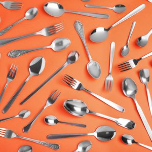 Silverware lies scattered across an orange background, consisting of various spoons and forks in different sizes and styles.
