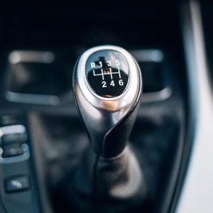 Gear shift knob displays transmission pattern in car interior, contrasting against dark surroundings. Text: R, 1, 2, 3, 4, 5, 6.