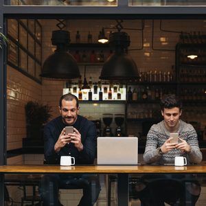 Two people sit using smartphones in a cozy cafÃ©, with a laptop and mugs on the wooden counter, surrounded by bottles and hanging lights.
