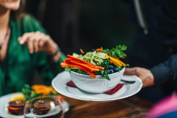 Horizontal image of male hand serving plate of delicious vegetarian mixed organic salad in restaurant to young smiling women. Waiter wearing black uniform.