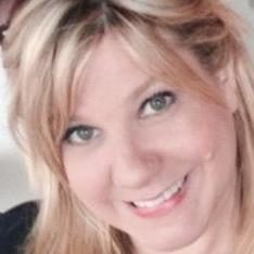 Smiling person with blonde hair looks forward, surrounded by a light, neutral background.