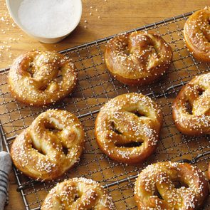 Golden pretzels rest on a cooling rack, sprinkled with coarse salt, beside a small bowl of additional salt on a wooden table.