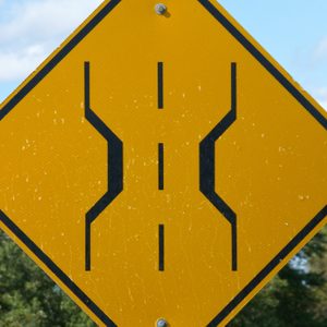 Yellow road sign indicates narrow bridge ahead, featuring black graphic lines, set against a background of green foliage and blue sky.