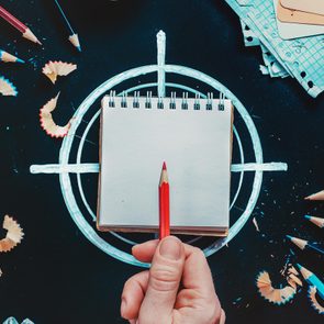 Hand holds red pencil over notepad, surrounded by pencil shavings, crumpled papers, and blue pencils on dark surface with drawn target.