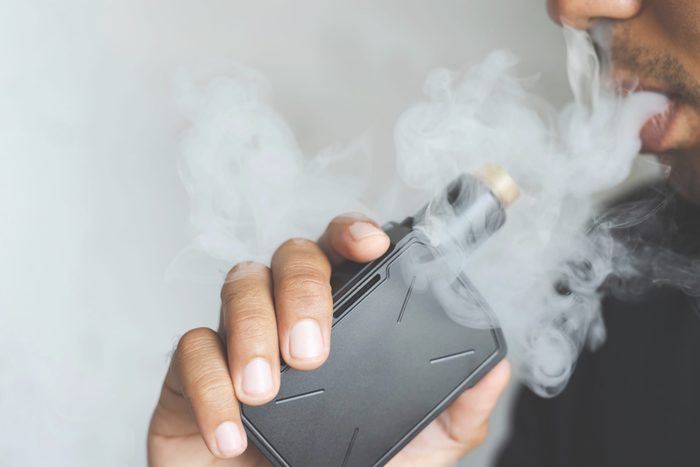 photo portrait young man with beard hold and smoking his electronic cigarette blowing a stream of smoke spread. dark black, grey background