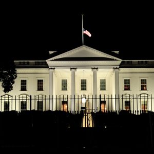 The White House illuminated at night, viewed through an iron fence, with an American flag flying atop, set against a dark sky.