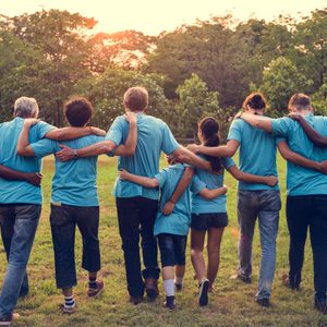 Group walks together with arms linked, wearing blue shirts, across a grassy field, surrounded by trees at sunset.