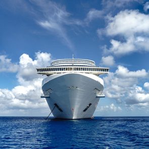 A large cruise ship sails forward, cutting through deep blue ocean, beneath a bright sky filled with scattered white clouds.