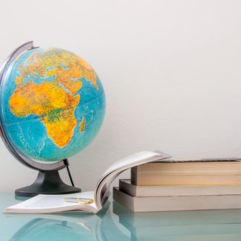 A globe rests on a table beside an open book and stacked books against a plain wall.
