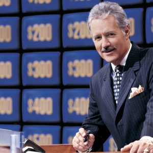 Host stands at a podium holding a pen, surrounded by a wall of screens displaying monetary values like 