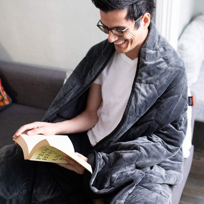 Person reading a book wrapped in a gray blanket, seated on a couch in a cozy room. The book's cover text: 