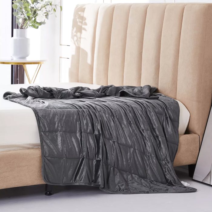 A dark gray blanket drapes over a bed with a beige upholstered headboard; a vase with flowers sits on a nearby side table.