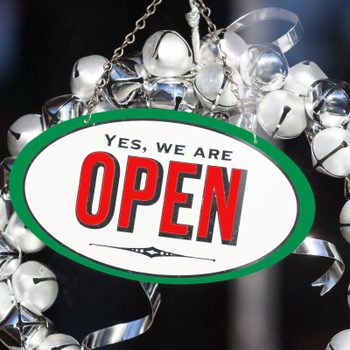 Sign hangs, reading "Yes, we are OPEN," surrounded by a wreath of shiny silver bells on a dark background.