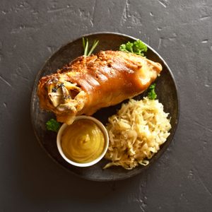 Roasted pork knuckle rests on a dark plate alongside sauerkraut and a bowl of mustard, garnished with parsley on a textured gray surface.