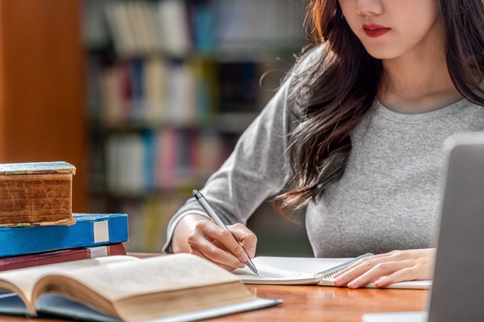 Closeup Asian young Student hand writing homework and using technology laptop in library of university or colleage with various book and stationary over the book shelf background, Back to school