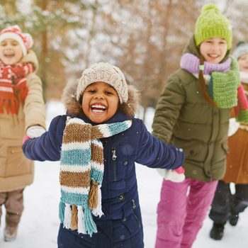 Children enjoy playing in snow, wearing colorful winter clothes. Trees and buildings surround them in the snowy, outdoor setting.