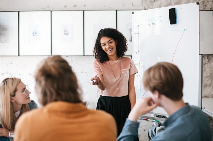 Beautiful African American lady with dark curly hair standing near board and happily discussing new project with her colleagues in office. Young smiling business woman giving presentation to coworkers