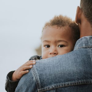 A small child rests on an adult's shoulder, gently holding them, wearing denim jackets in an outdoor setting.