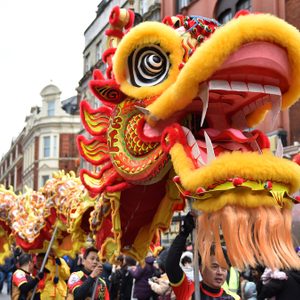 Dragon puppet parades through city street, carried by performers, surrounded by onlookers and buildings, celebrating a festive occasion with vibrant colors and decorations.
