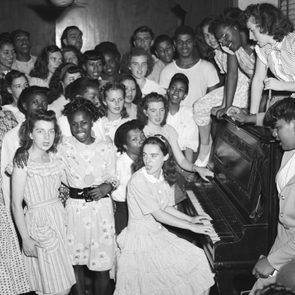 A group of people sings around a piano, where a young woman plays. They are in a lively social gathering indoors.