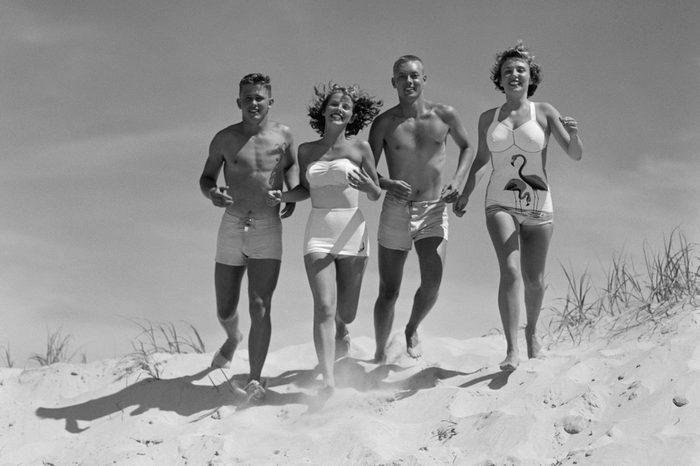 1950s TEENAGE FOURSOME TWO COUPLES IN BATHING SUITS RUNNING ON BEACH TOWARD OCEAN