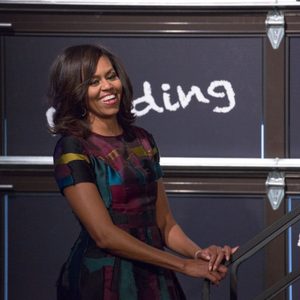 Woman smiles standing beside railing; wears colorful dress. Background features dark wall with white text: 