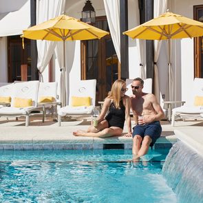 Couple relaxes by pool, holding drinks, surrounded by yellow umbrellas and white lounge chairs at a sunny resort.
