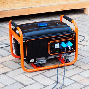 A portable generator sits with cables plugged in, surrounded by a paved surface, near a wooden structure.