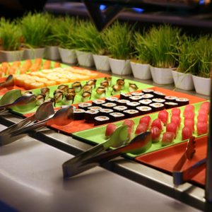 Sushi displayed on colorful trays with metal tongs, surrounded by decorative pots of grass, inside a buffet-style setting.