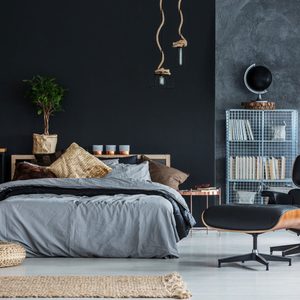 Bed with gray bedding sits against a dark wall, surrounded by hanging lights, a plant, bookshelves, and a lounge chair in a modern bedroom.