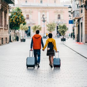 Two people holding hands, pulling suitcases down a cobblestone street, surrounded by historic buildings and potted trees.