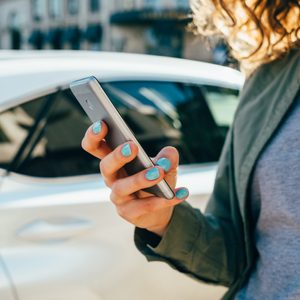 Person holding a smartphone, standing beside a white car on a city street. Brightly painted blue nails, wearing a gray shirt and necklace.