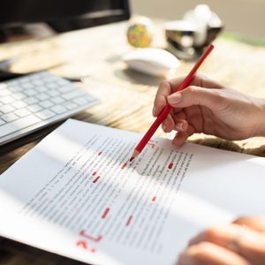 Hand editing a document using a red pencil on a wooden desk, next to a keyboard and monitor, in a sunlit workspace.