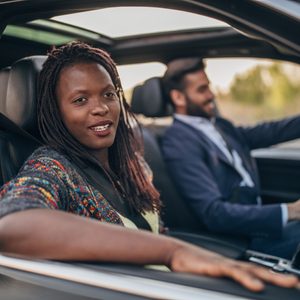 Two people sit in a car, smiling. The context is an outdoor setting with sunlight filtering through the trees.