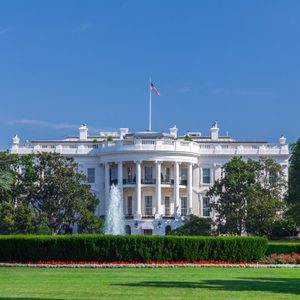 White building stands prominently with a flag flying, surrounded by lush gardens and trees under a clear blue sky.