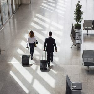 Two people pull rolling suitcases while walking through a spacious, sunlit airport terminal with empty seating areas and a potted plant nearby.