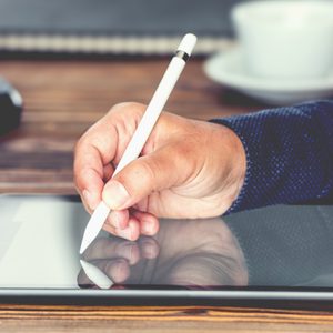 A hand holds a stylus, writing on a tablet in a wooden desk setting with a keyboard, mouse, and cup in the background.