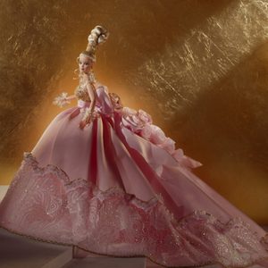 Doll elegantly poses, wearing an opulent pink gown adorned with floral lace and embellishments, against a textured gold backdrop.