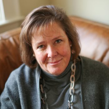 Woman smiling, sitting on a leather sofa, wearing a gray top and chain necklace, in a softly lit room with beige walls.