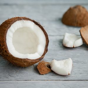 A halved coconut rests open, exposing white flesh, with scattered shell pieces on a weathered wooden surface.