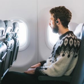 Person wearing earbuds, sitting and looking out an airplane window, surrounded by rows of empty seats.