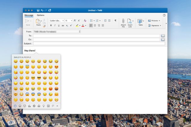 Email draft with emoji panel open displays various smiley faces; background shows a cityscape from above under a clear blue sky.