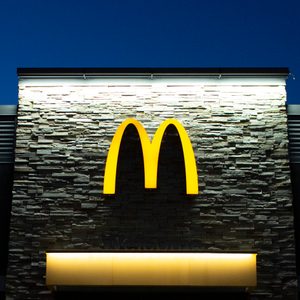 Yellow logo glows on stone wall, representing a fast-food restaurant, under a darkening sky. Text below states 