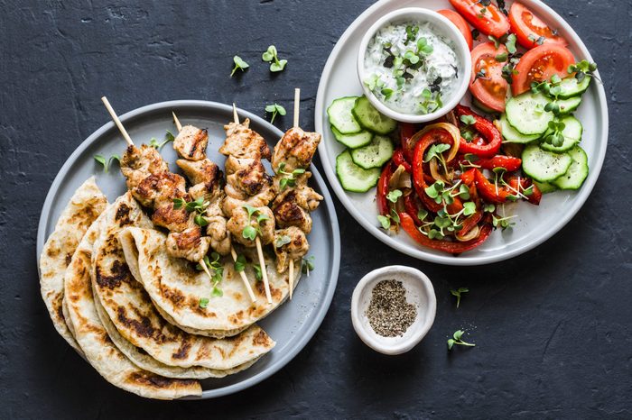 Mediterranean style lunch table - turkey skewers, flatbread, tomatoes, cucumber salad, baked sweet pepper, yogurt herb sauce on a dark background, top view