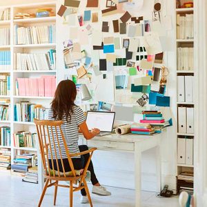 Person using a laptop, sitting at a cluttered desk, surrounded by books and colorful papers pinned on the wall in a home office.
