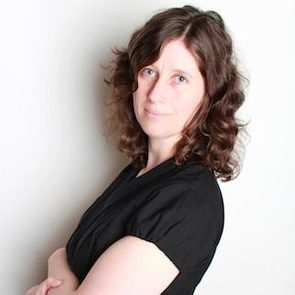 A person with curly hair stands facing the camera, arms crossed, against a plain white background.