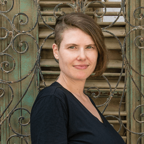 A person stands smiling, wearing a black shirt, in front of an ornate, teal-colored metal lattice with wooden shutters.