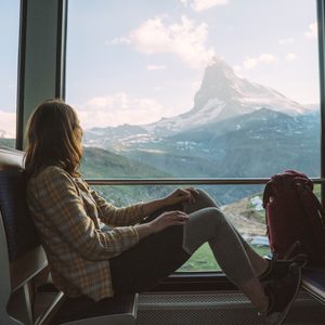 A person sits in a train, watching a scenic mountain landscape through the window, with a backpack beside them.